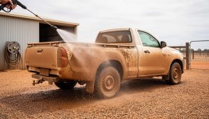 Dusty farm ute being pressure washed with a hose wand, with mist cleaning grime from the wheel arch and side panel